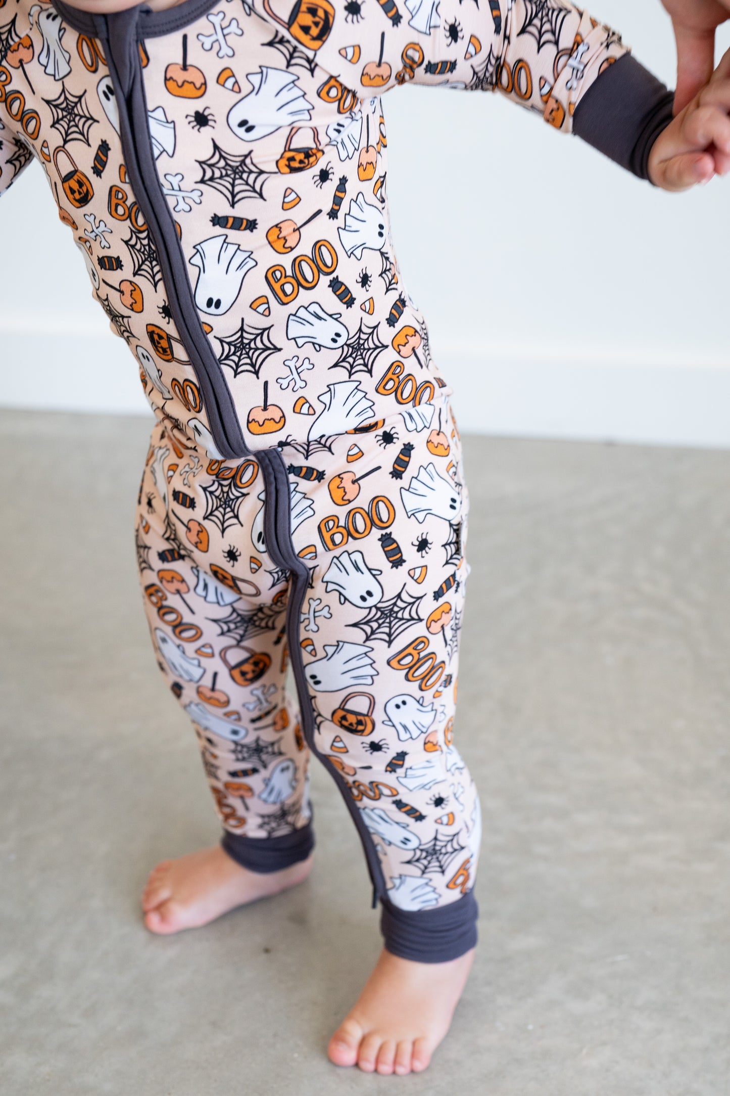 Child wearing Halloween-themed onesie with pumpkins, ghosts, and spider webs on a neutral background.